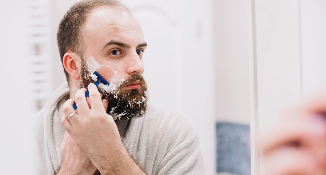 Homem em frente ao espelho usando aparelho de barbear para fazer degradê na barba em casa.